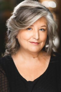 White Woman With Shoulder Length Wavy Grey Hair Wearing A Scoop Necked Black Shirt.