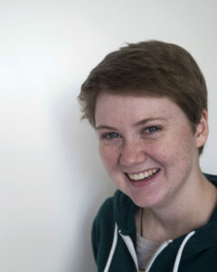 White Woman With Short Reddish Blond Hair Wearing A Black Hoodie. Smiling Against A White Background.