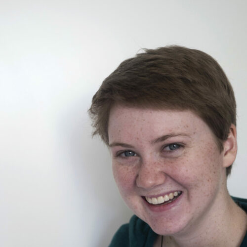 White Woman With Short Reddish Blond Hair Wearing A Black Hoodie. Smiling Against A White Background.