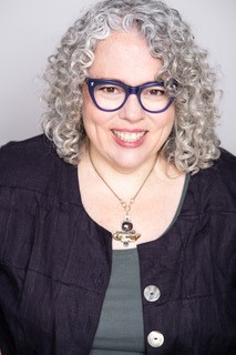 White woman with black rimmed glasses and curly grey shoulder length hair wearing a grey top and black cardigan with a large pendant necklace