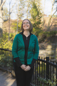 White Woman With Chin Length Straight Brown Hair Wearing Black Pants And Shirt With A Green Jacket, Standing Outside In A Park Smiling At The Sky