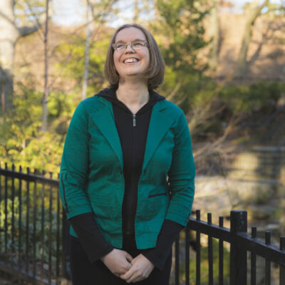 White Woman With Chin Length Straight Brown Hair Wearing Black Pants And Shirt With A Green Jacket, Standing Outside In A Park Smiling At The Sky