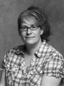 White Woman With Glasses And Light Brown Hair Swept Back In A Pony Tail Wearing A Plaid Short Sleeved Shirt Against A Grey Background.