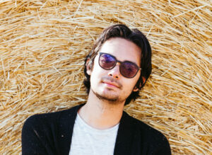 Filipino Man With Dark Short Hair And Goatee Laying On A Stack Of Hay Wearing Sunglasses, Grey T-shirt And Black Blazer