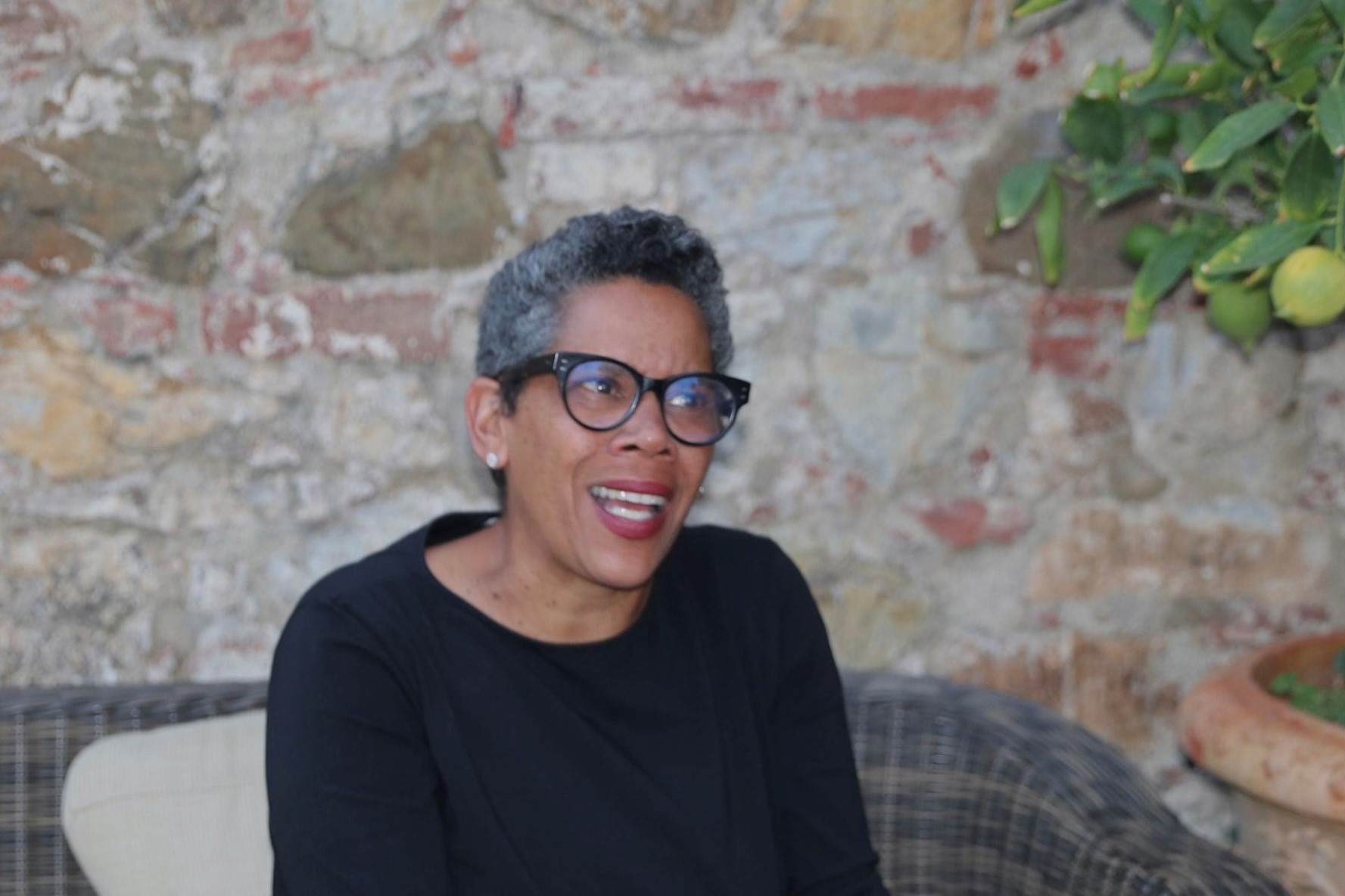 Black woman with greying dark hair wearing a long sleeved black top and thick black framed glasses in front of a stone wall